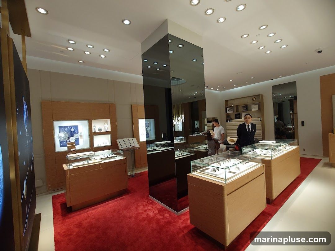 The interior of a high-end watch store with polished wooden display cases, glass counters showcasing watches, and a red carpet. Two staff members are visible, one in a suit and tie, and another in casual attire. The store is well-lit with recessed ceiling lights.