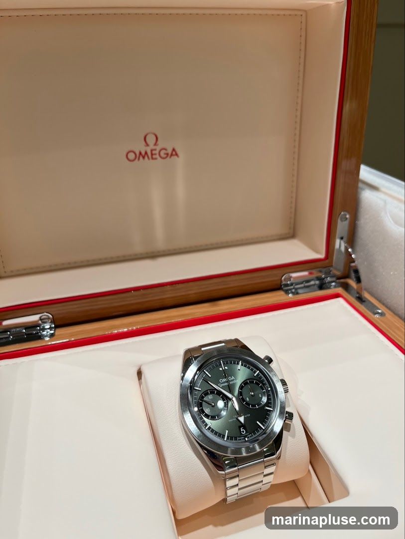 A close-up view of an Omega watch with a green dial, presented in its open wooden box. The Omega logo is visible in red on the inside lid of the box.
