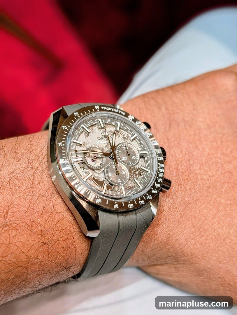 A close-up shot of an Omega Speedmaster watch on a person's wrist. The watch has a grey dial with intricate detailing and sub-dials. The bezel features tachymetre markings. The watch is fitted with a grey rubber strap.