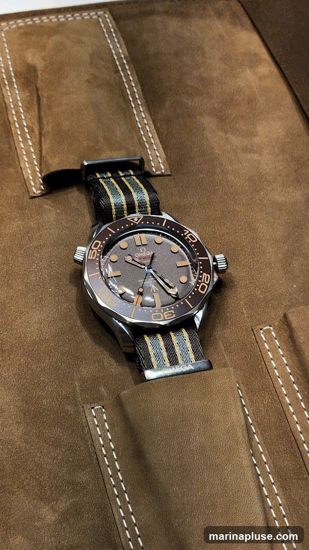 A close-up shot of an Omega Seamaster watch with a brown dial and a striped NATO strap. The watch is resting on a brown suede-like material with visible stitching. The Omega logo is visible on the dial and on the watch strap buckle.