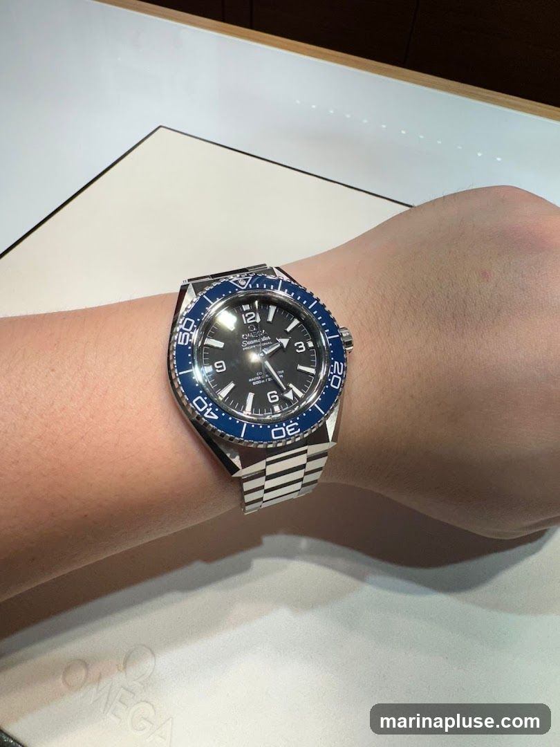 A close-up shot of a person's wrist wearing a black-faced Omega Seamaster watch with a blue bezel and a stainless steel bracelet. The watch is displayed on a white surface with the embossed Omega logo visible in the lower portion of the image.