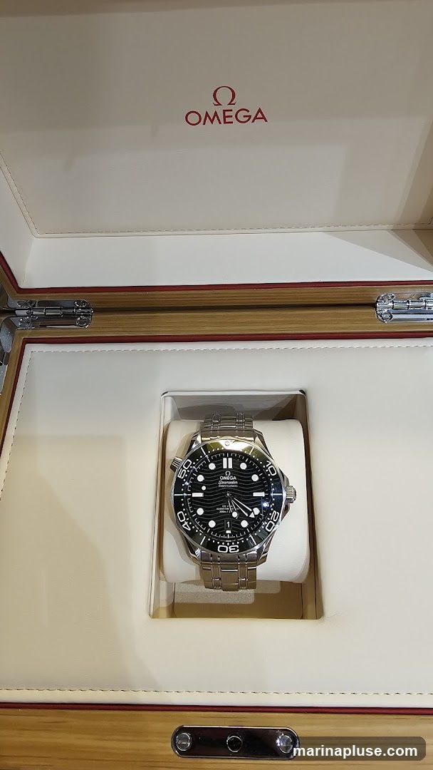 An Omega Seamaster watch with a black dial and stainless steel bracelet is displayed inside its presentation box. The box is lined with cream-colored material and features the red Omega logo and name on the lid. The watch is resting on a cream-colored cushion within the box.