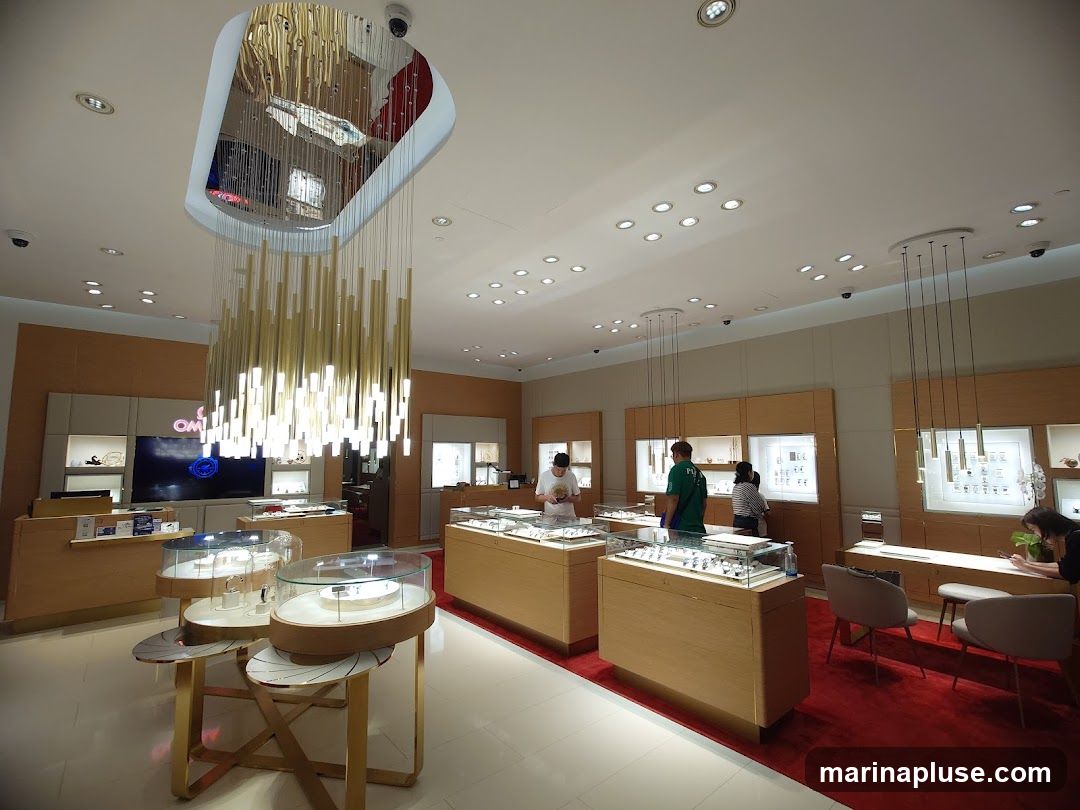 The interior of a modern watch store with display cases filled with watches. A large, ornate chandelier hangs from the ceiling. Several customers and staff members are visible browsing the merchandise. The store features wooden display cabinets and red carpeting. A large screen displays the OMEGA logo.