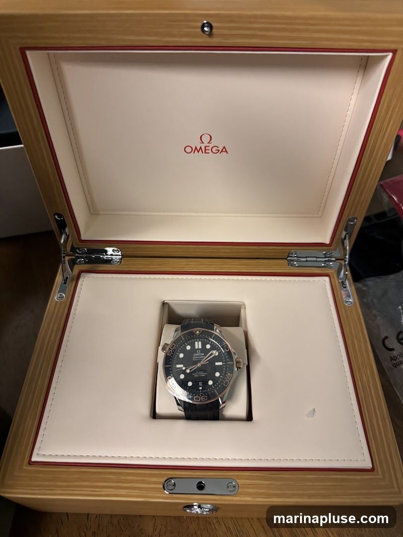 An open wooden box with the OMEGA logo in red on the inside lid. A black-strapped Omega watch with a dark dial and rose gold accents is displayed in a cutout within the box.