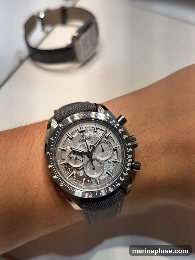 A close-up shot of an Omega Speedmaster watch on a person's wrist. The watch has a grey dial with three subdials and a tachymetre bezel. In the blurred background, another watch with a black strap is visible.