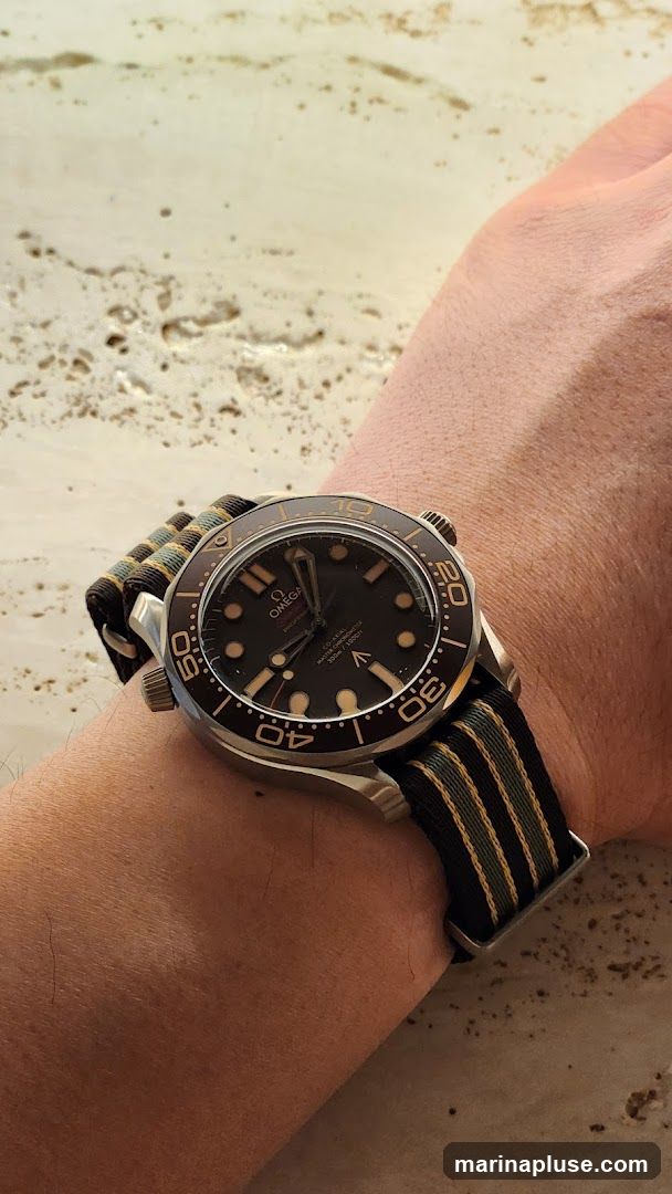 A close-up shot of an Omega Seamaster watch on a person's wrist. The watch has a black dial with luminous hour markers and hands. The bezel is brown and has gold markings. The watch is on a striped NATO strap in black, green, and gold. The background appears to be a sandy or textured surface.
