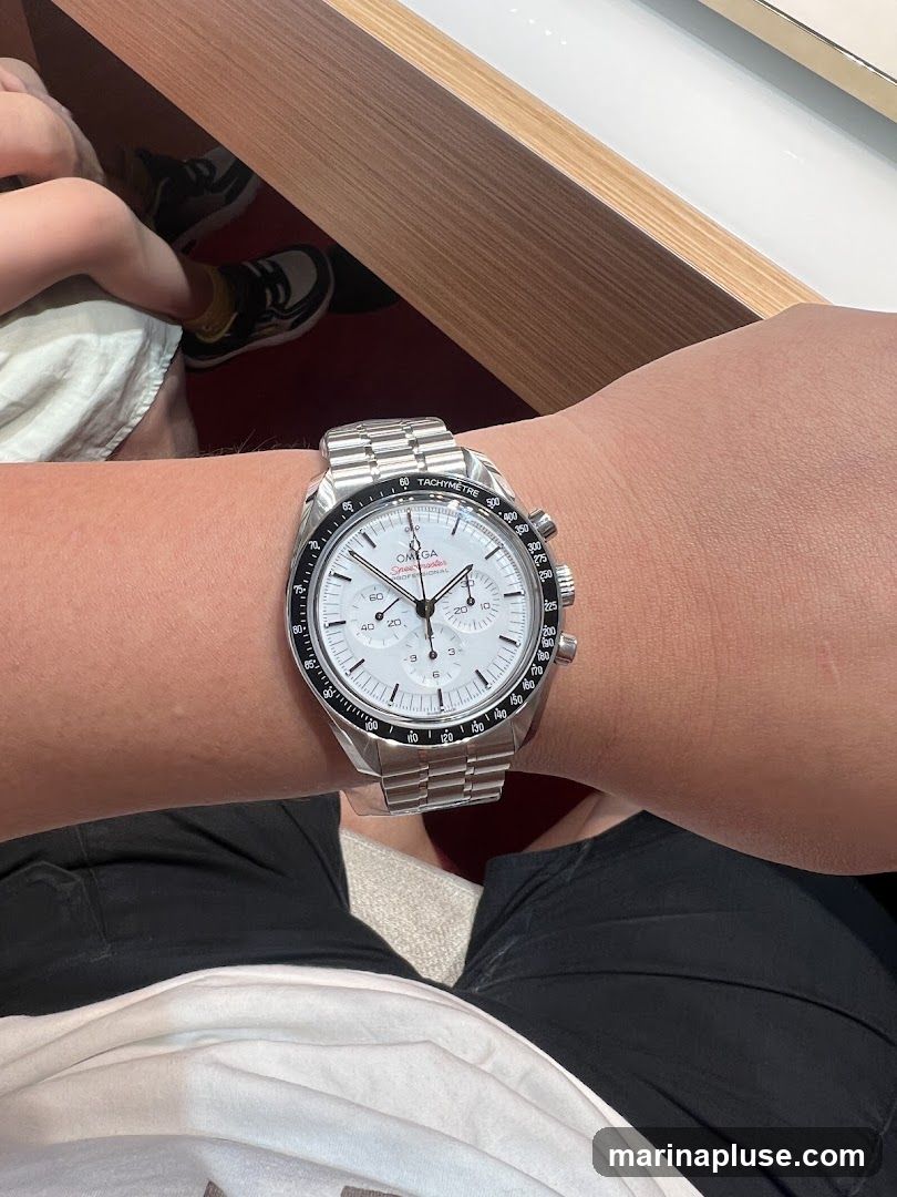 A close-up shot of a person wearing a silver Omega Speedmaster Professional watch on their left wrist. The watch has a white dial with black markers and hands, and a black tachymetre bezel. The watch is on a metal bracelet. In the background, a child's legs and feet are visible, wearing sneakers and socks. The setting appears to be indoors, possibly a retail environment.