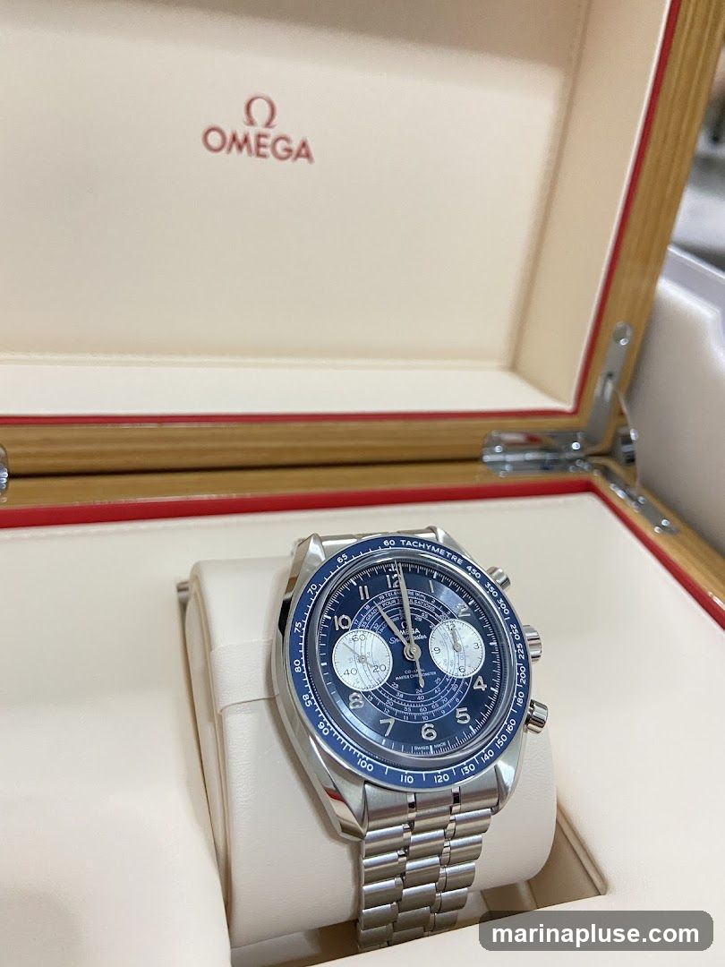 A close-up shot of an Omega watch with a blue dial and silver band, resting inside its presentation box. The Omega logo is visible on the inside lid of the box.