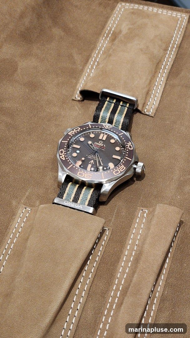 A close-up shot of an Omega Seamaster watch with a striped NATO strap, resting on a tan suede-like material with stitched detailing. The watch face is dark with luminous hour markers and hands. The bezel is also visible, with markings for minutes. The OMEGA logo is visible on the watch face and the strap.
