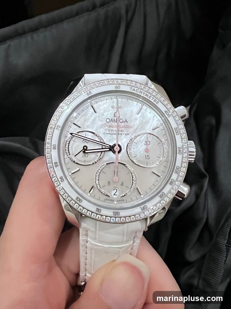 A close-up shot of a white Omega Speedmaster watch with a diamond-encrusted bezel and a white alligator leather strap. The watch face is mother-of-pearl with rose gold accents and sub-dials. The watch is held by a person's hand against a dark, textured background.