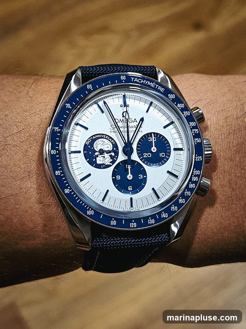A close-up shot of an Omega Speedmaster watch on a person's wrist. The watch has a white dial with blue accents and a blue bezel. It features a tachymeter scale and sub-dials. A graphic of Snoopy with the text "50TH ANNIVERSARY" is visible on the dial. The watch is worn with a dark blue strap.