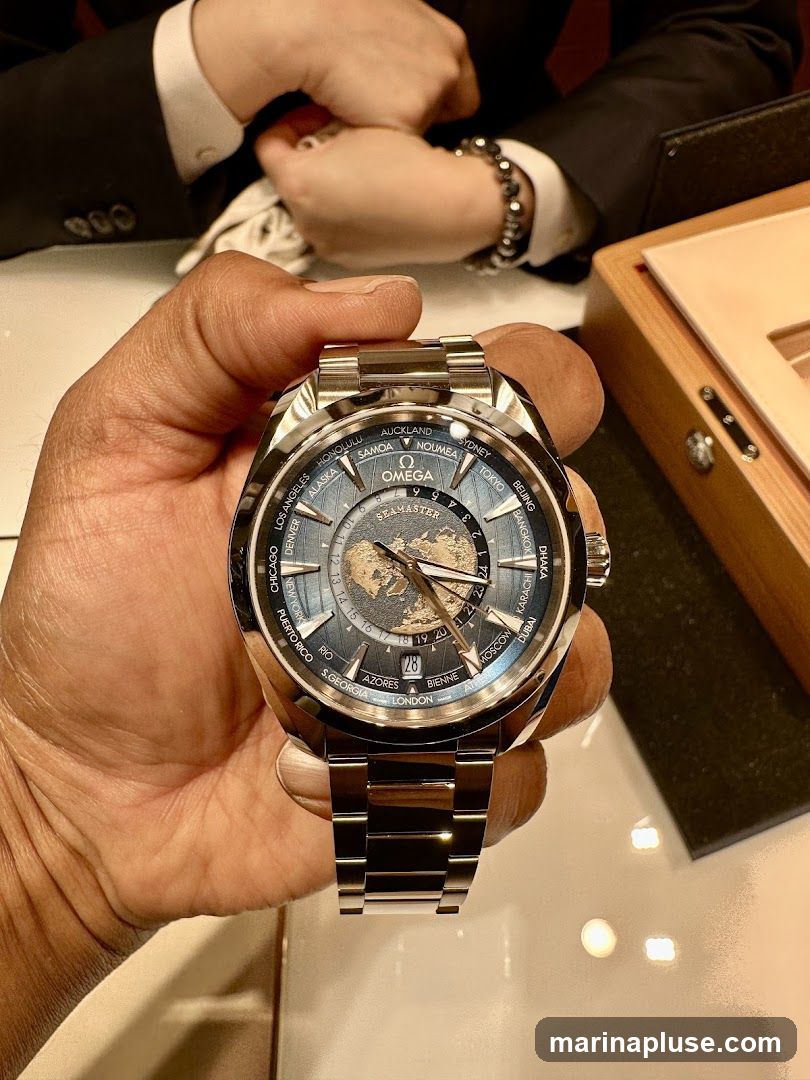 A close-up shot of a person holding an Omega Seamaster watch with a world time complication. The watch has a blue dial with a depiction of the Earth and city names around the bezel. In the background, two people in suits are visible, one wearing a bracelet. A wooden watch box is also partially visible.