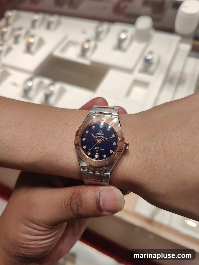 A close-up shot of a person wearing an Omega Constellation watch on their wrist. The watch has a dark blue dial with diamond hour markers and Roman numerals on the bezel. The watch band is a combination of silver and rose gold. In the blurred background, several other watches are displayed on white stands in a jewelry store or watch shop.