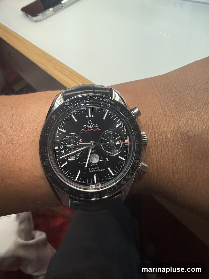 A close-up shot of an Omega Speedmaster watch on a person's wrist. The watch has a black dial with silver hands and markers. It features a tachymetre scale on the bezel, sub-dials for chronograph functions, and a moonphase complication. The text "OMEGA Speedmaster", "CO-AXIAL MASTER CHRONOMETER", and "SWISS MADE" are visible on the dial.