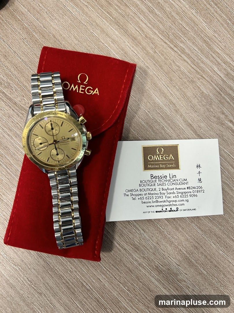 A gold and silver Omega Speedmaster watch is displayed on a red Omega pouch. Next to it is a business card for Bessie Lin, a Boutique Technician and Sales Consultant at the Omega Boutique in Marina Bay Sands, Singapore. The card includes contact information and the Omega logo.