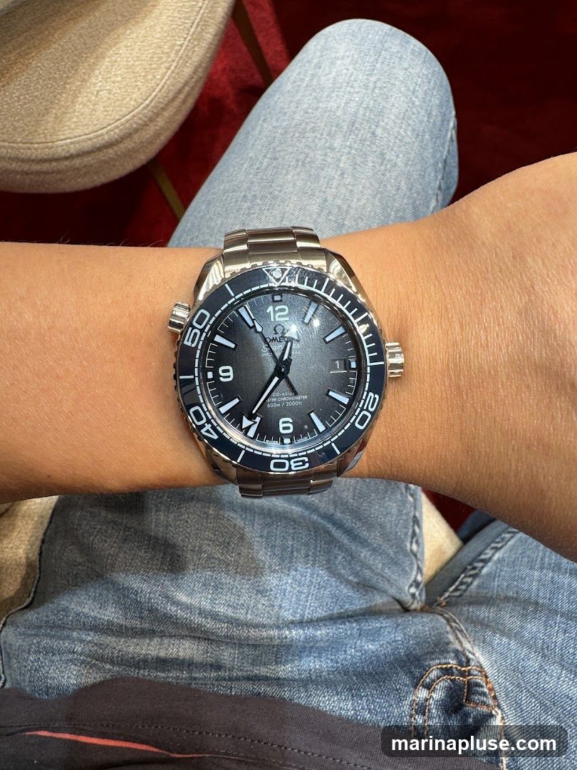 A close-up shot of a person wearing a silver Omega Seamaster watch on their wrist. The watch has a dark grey dial with silver hour markers and hands. The bezel is black with white numerals. The person is wearing blue jeans.