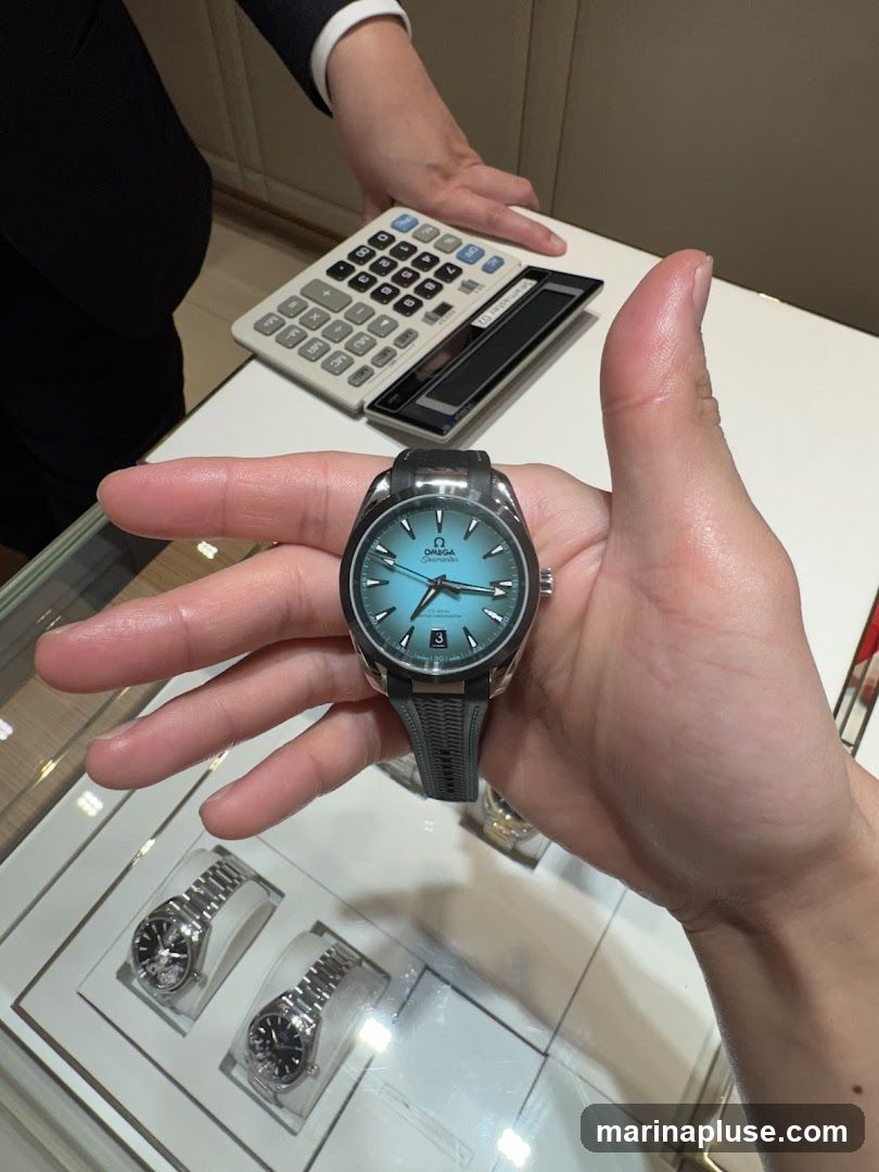 A person is holding a blue-faced Omega watch with a black strap. In the background, a calculator and other watches are visible on a display counter. A person in a suit is partially visible in the upper left corner.