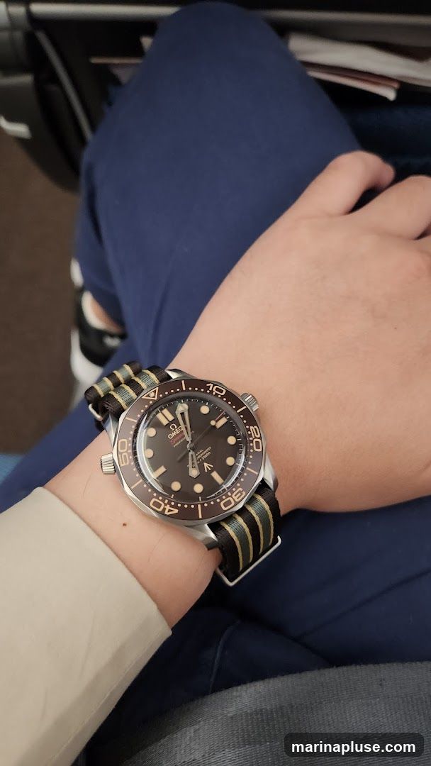 A close-up shot of a person's wrist wearing a watch with a dark dial and a striped NATO strap. The watch has a rotating bezel with minute markers. The person is wearing a light-colored shirt and dark blue pants.