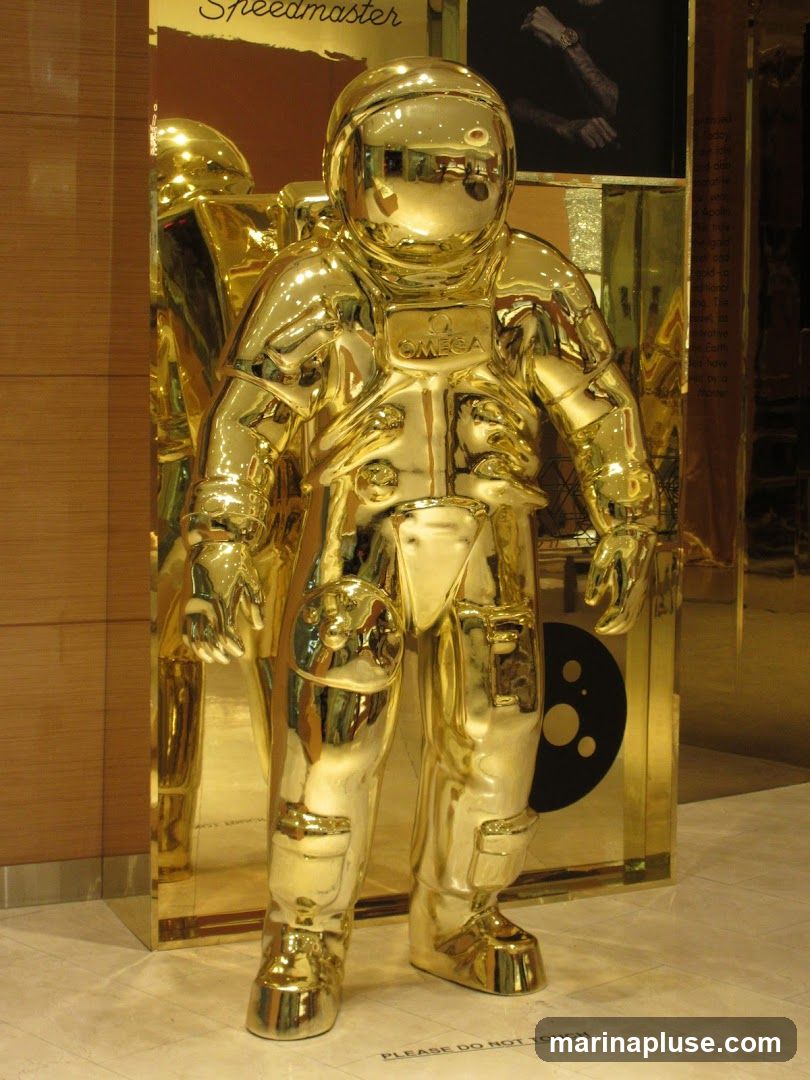 A full-body, shiny golden statue of an astronaut stands in a display. The astronaut is wearing a helmet and a detailed spacesuit. The Omega logo is visible on the chest plate. Behind the astronaut, there is a reflective golden panel and a dark circular graphic with several smaller circles. Text above the astronaut reads "Speedmaster". A sign on the floor says "PLEASE DO NOT TOUCH".