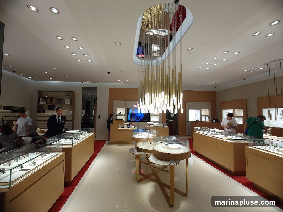 The interior of a high-end watch store with multiple display cases filled with watches. Customers and staff are browsing the merchandise. A large, modern chandelier hangs from the ceiling. The store appears well-lit and spacious.