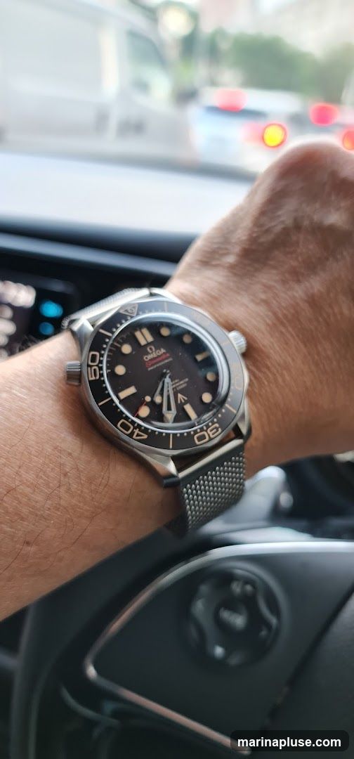 A close-up shot of a person's wrist wearing an Omega Seamaster Professional watch. The watch has a black dial with silver markers and hands, and a rotating bezel with minute markers. The watch is on a mesh metal strap. The background is blurred, showing what appears to be a car interior with traffic outside.