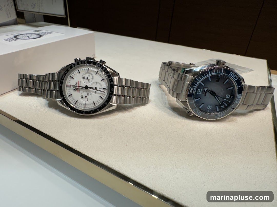 Two Omega brand watches are displayed on a white surface. The watch on the left has a white dial with three sub-dials and a tachymeter bezel. The watch on the right has a dark grey dial with a rotating bezel. A white box with an Omega logo is visible behind the left watch.