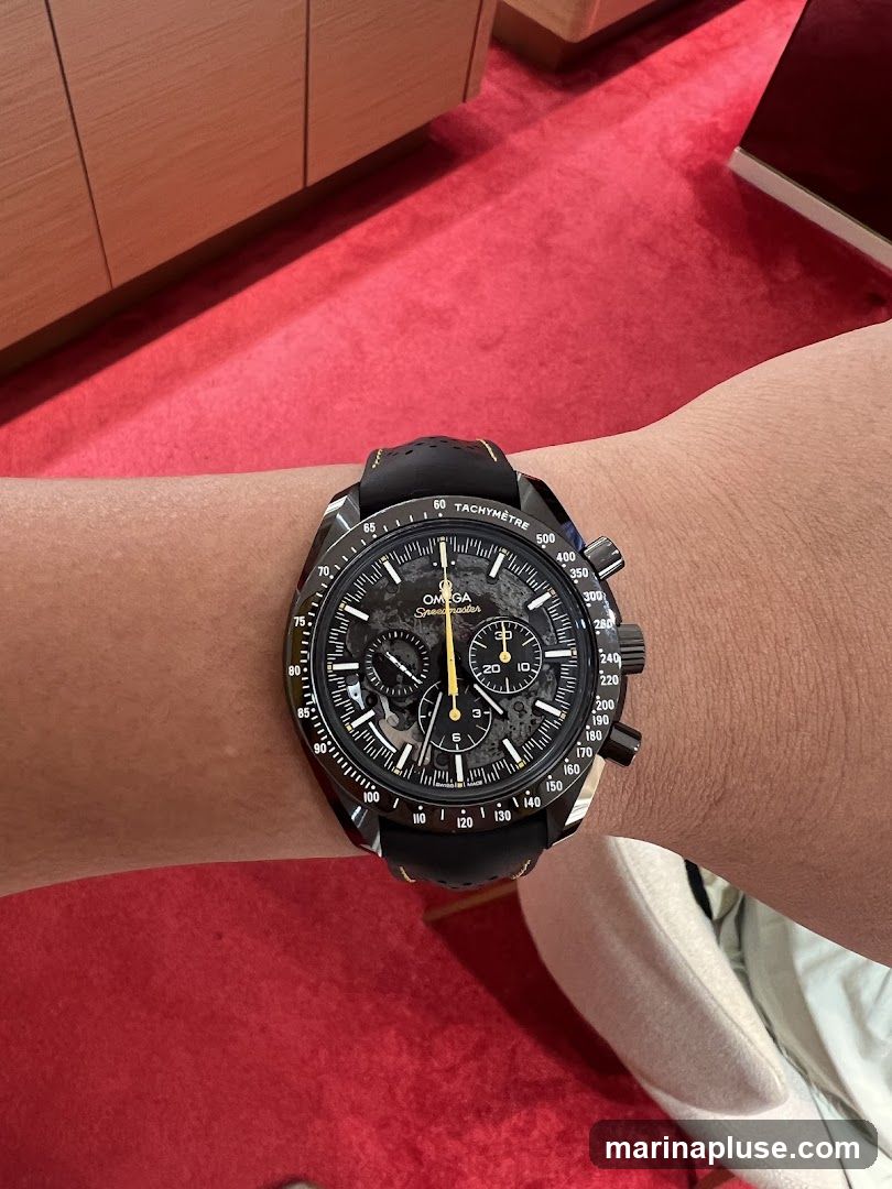 A close-up shot of a person wearing a black Omega Speedmaster watch on their wrist. The watch has a black dial with yellow accents and a black leather strap. The background shows a red carpet and wooden cabinetry.