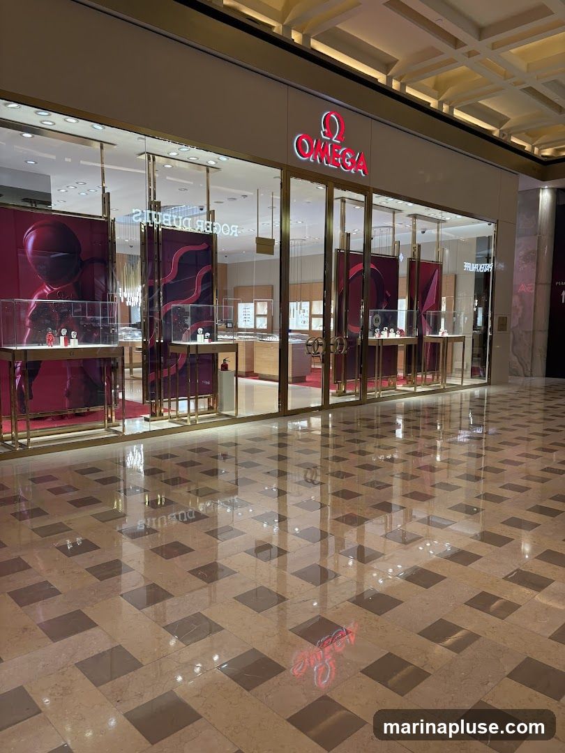 The exterior of an Omega watch store. The store has a large glass facade with gold trim. The Omega logo and name are prominently displayed above the entrance. Inside, display cases showcase watches, and there are large red banners with abstract designs. The floor is polished marble with a checkered pattern.