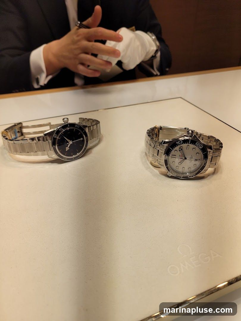 Two luxury Omega watches are displayed on a white surface. The watch on the left has a black dial and a stainless steel band. The watch on the right has a white dial with black accents on the bezel and a stainless steel band. A person in a suit and white gloves is visible in the background, appearing to present or discuss the watches. The Omega logo is embossed on the display surface.