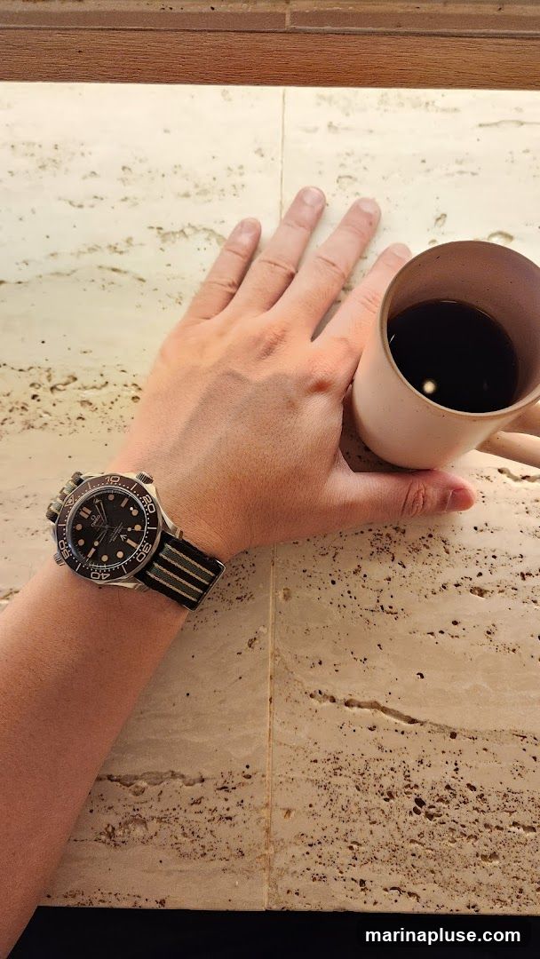 A person's hand is holding a light-colored mug filled with dark coffee. The person is also wearing a watch with a black dial and a striped band on their wrist. The background appears to be a textured, light-colored surface, possibly stone or tile, with darker speckles. The image is taken from a top-down perspective.
