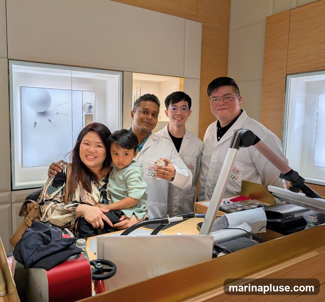 A family of three, a woman holding a young child, poses with three men in white lab coats. The men appear to be watchmakers, as one is holding a watch and the lab coats have the OMEGA logo and Olympic rings. They are standing behind a counter in what appears to be a watch store, with display cases visible in the background.