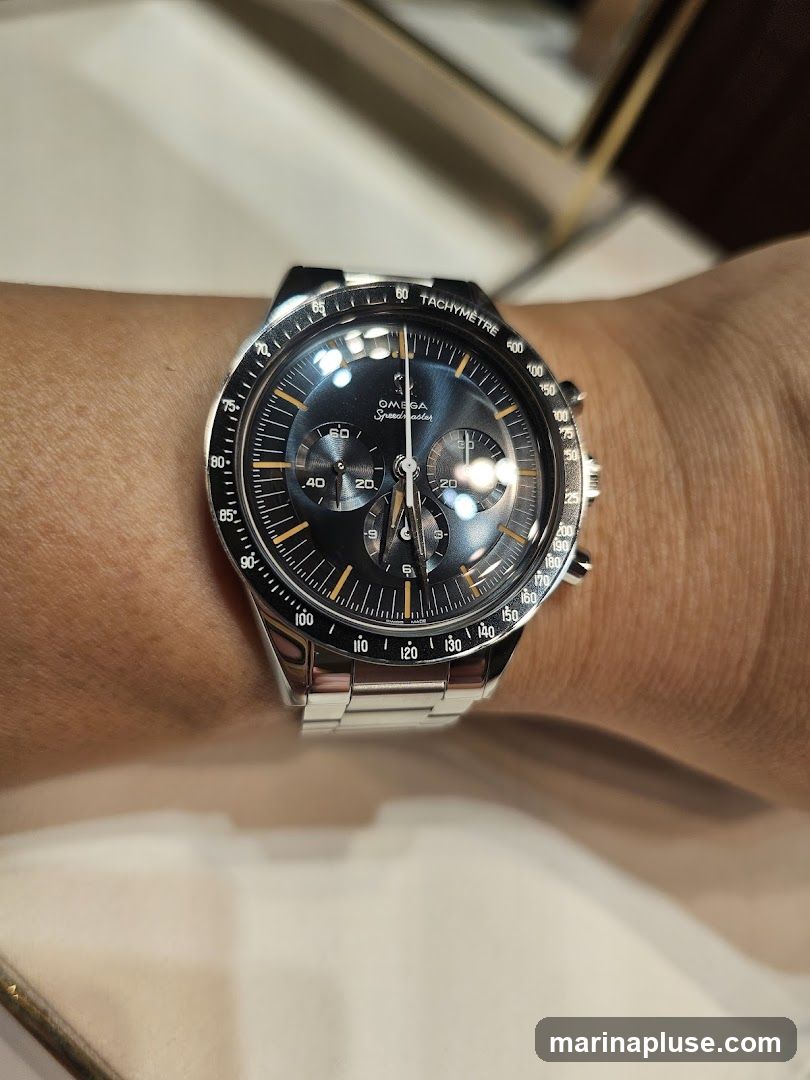 A close-up shot of an Omega Speedmaster watch on a person's wrist. The watch has a dark dial with silver hands and markers, and a tachymeter bezel. The metal bracelet is also visible.