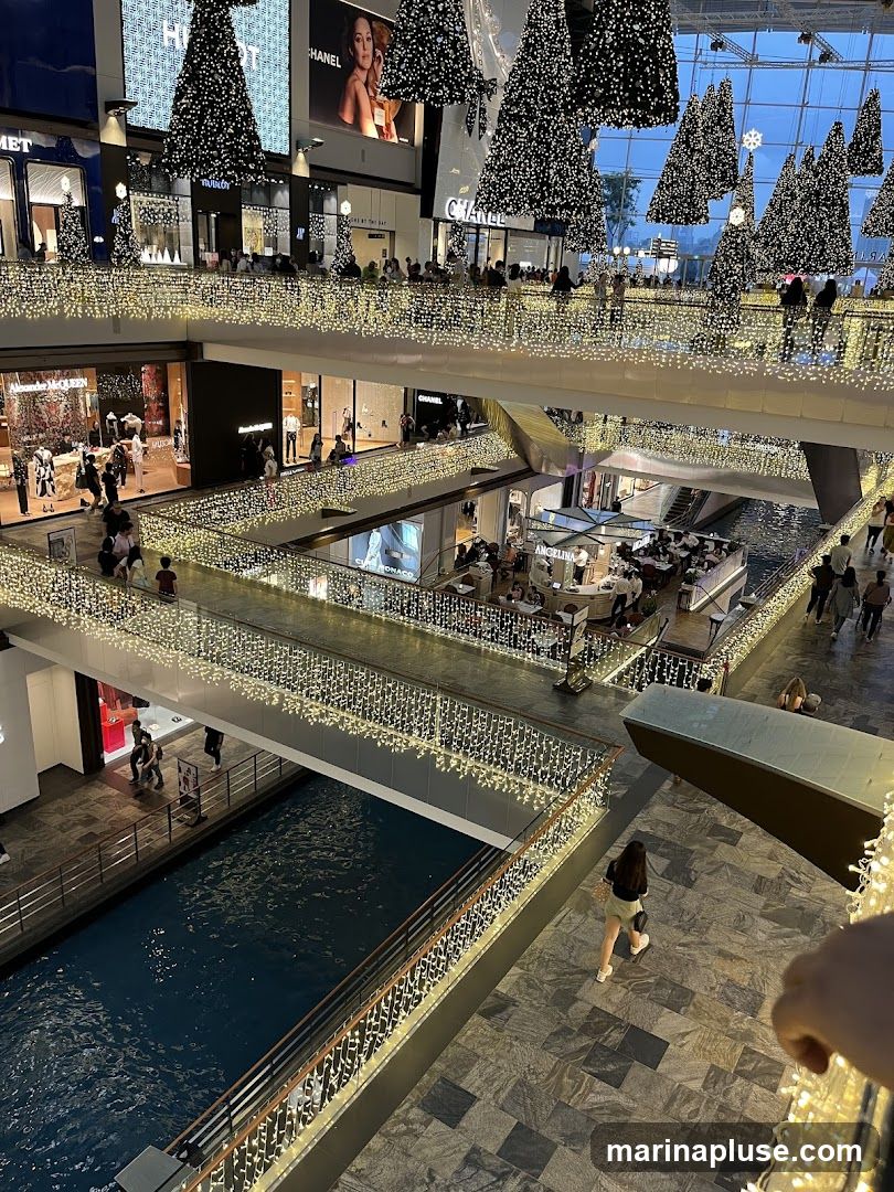 An interior view of a multi-level shopping mall decorated for Christmas. Numerous artificial Christmas trees adorned with lights are suspended from the ceiling. Balconies and walkways are lined with strings of warm white fairy lights. People are seen browsing shops and walking along the elevated pathways. A canal with blue water runs through the lower level of the mall. Shops visible include Chanel and Alexander McQueen.