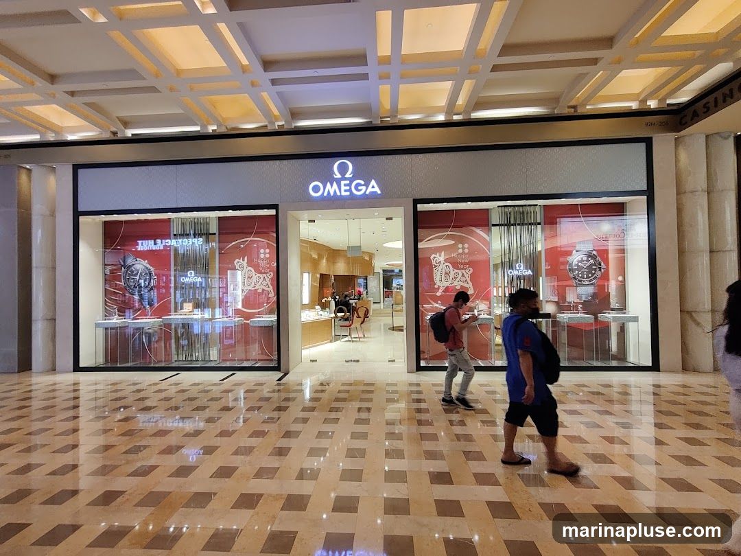 The exterior of an Omega watch store with large display windows showcasing watches. The Omega logo is prominently displayed above the entrance. People are walking by the storefront.