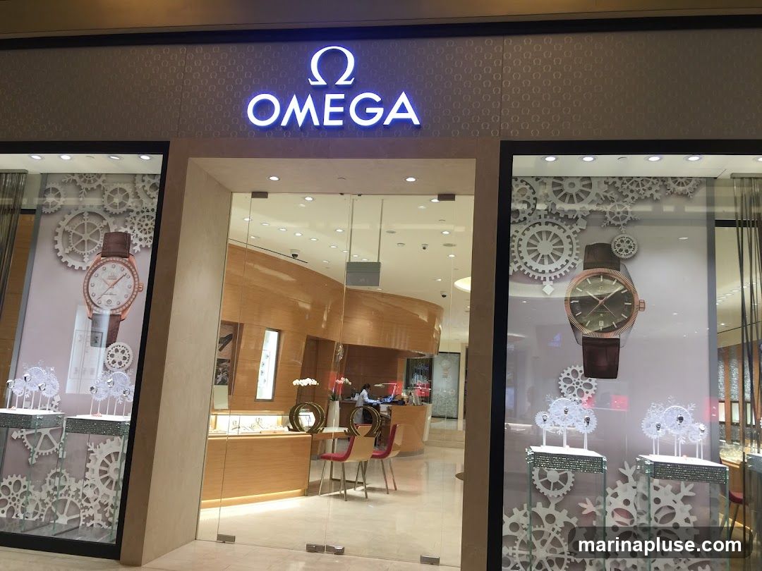 The exterior of an Omega watch store. The store has a large glass entrance and display windows on either side. The windows feature large images of watches and decorative gear motifs. The Omega logo and name are prominently displayed in illuminated lettering above the entrance. Inside, the store appears modern and well-lit, with display cases and seating areas visible.