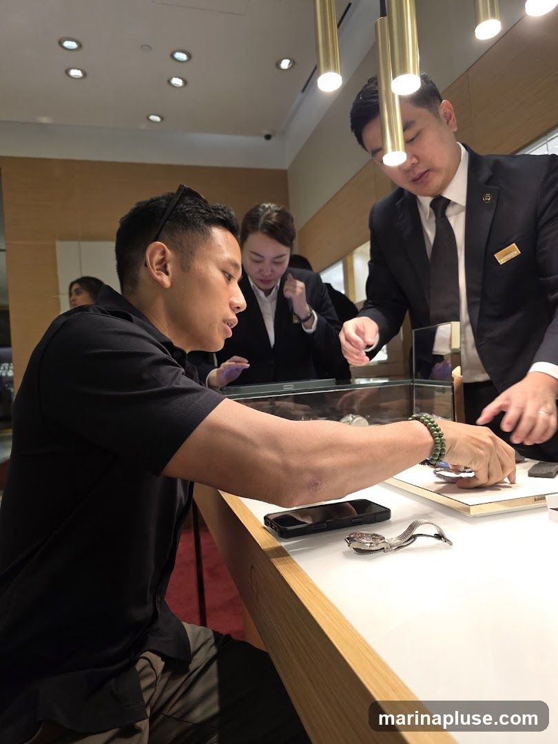 A man in a black polo shirt is seated at a counter, looking at a watch. Two sales associates in suits are standing behind the counter, assisting him. One associate is showing him something on a tablet, and the other is holding a watch. There is a watch on the counter in front of the customer. The store appears to be a high-end jewelry or watch store.