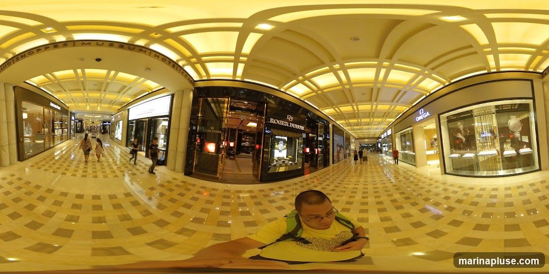 A wide-angle view of a brightly lit, upscale shopping mall interior. The floor is paved with a checkered marble pattern. Shops line both sides of the walkway, featuring luxury brands like Roger Dubuis and Omega, with their storefronts displaying watches and jewelry. Several shoppers are visible walking through the mall. A person wearing a yellow shirt and backpack is in the foreground, looking down at their phone. The ceiling has an intricate, illuminated design. Signs indicate "SHOPPES | MUSEUM | HOTEL".