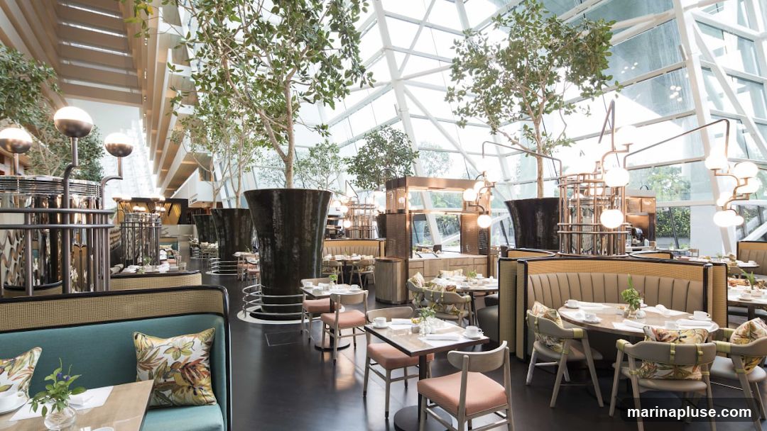 A spacious and modern restaurant interior featuring a high ceiling with a geodesic glass roof, allowing ample natural light. Several large potted trees are strategically placed throughout the dining area, adding a touch of nature and creating a serene ambiance. The seating arrangements include comfortable booths with plush upholstery and elegant chairs, all set around tables adorned with white tablecloths, place settings, and small floral arrangements. The decor is sophisticated, with warm lighting fixtures and a neutral color palette accented by the vibrant green of the plants and the colorful cushions on some of the chairs.