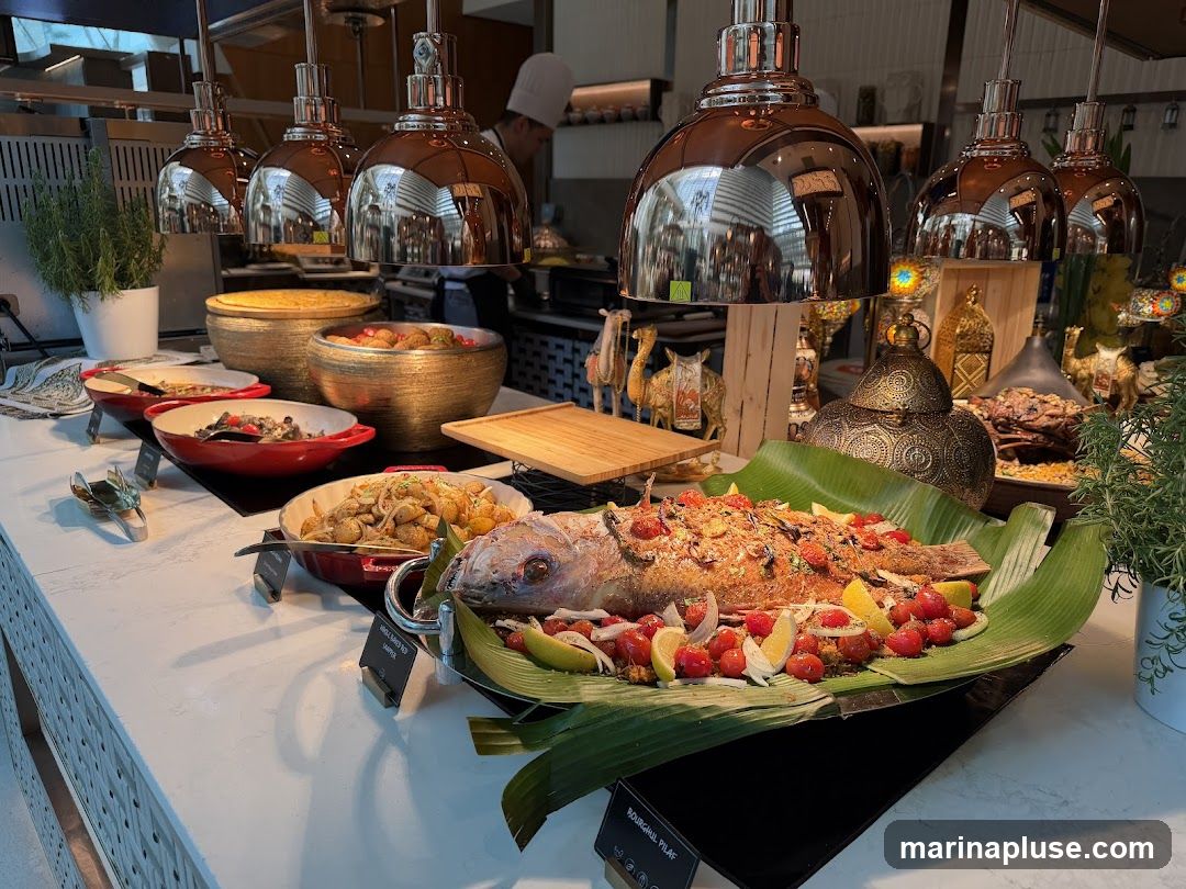 A buffet table is laden with various dishes, prominently featuring a whole roasted fish garnished with cherry tomatoes, lemon slices, and onions, resting on a bed of banana leaves. Several chafing dishes and bowls contain other food items, including what appears to be potatoes and other mixed dishes. Decorative elements such as ornate lanterns and camel figurines add to the ambiance. A chef is visible in the background, working in the kitchen area.