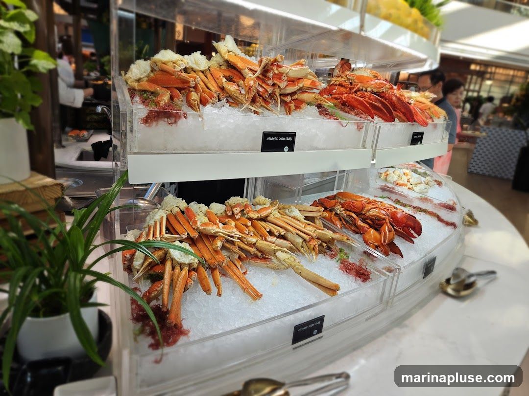 A close-up view of a seafood buffet display featuring an abundance of crab legs and lobsters on ice. The display is well-lit, highlighting the vibrant colors of the seafood. In the background, blurred figures of people and other food items suggest a busy dining environment. Small labels are visible on some of the containers, identifying the seafood.