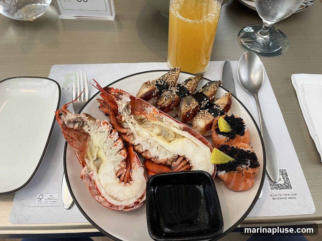 A plate of food containing a lobster tail, sushi rolls topped with black caviar and lime, and several pieces of eel sushi. A glass of orange juice and a glass of water are also visible on the table. A QR code on the placemat suggests scanning for a beverage menu.