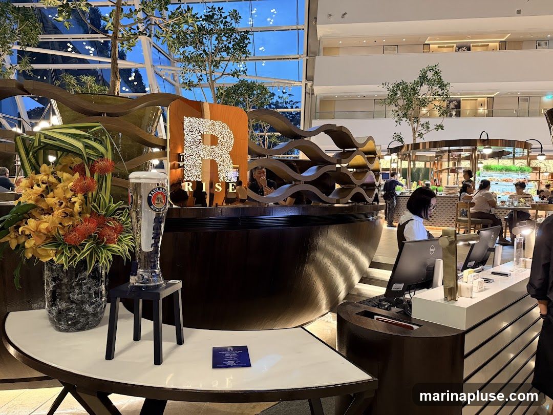 The image shows the interior of a restaurant with a prominent "RISE" sign featuring a large decorative "R". In the foreground, there is a tall beer glass on a small stand next to a vase of yellow and orange flowers. The background reveals a buffet area with people serving themselves and seated at tables. The overall ambiance suggests a modern and upscale dining establishment with natural elements like plants and a glass ceiling.