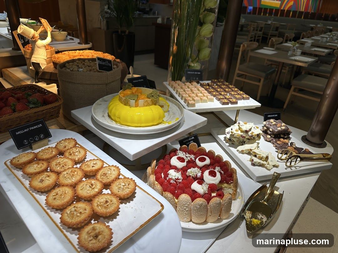 A dessert buffet with various sweet treats. In the foreground, there are small hazelnut financiers on a white platter. To the right, a raspberry and ladyfinger cake is displayed. Behind these, a yellow cake with fruit and pistachios sits on a tiered stand. Further back, rows of small chocolates and white chocolate bark with dried fruit and nuts are arranged. A basket of strawberries is visible on the left. The background shows a dining area with tables and chairs.