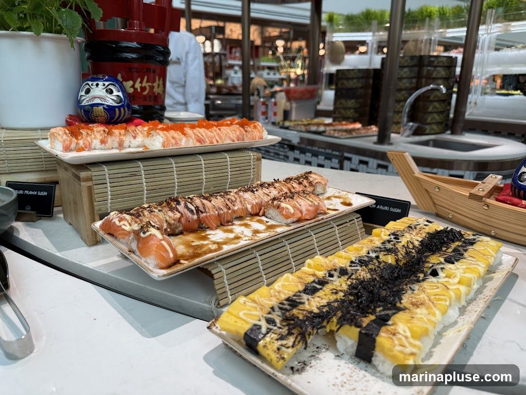 A close-up view of a sushi buffet with several platters of sushi. In the foreground, there are two platters of sushi rolls. The one on the right is topped with yellow egg, mayonnaise, and seaweed. The one on the left features salmon with a drizzle of sauce and mayonnaise. Behind these, a platter of sushi rolls topped with orange roe and shrimp is visible. A decorative Daruma doll sits atop this platter. In the background, more sushi and other food items are displayed on a buffet line, with a person in a white uniform visible.