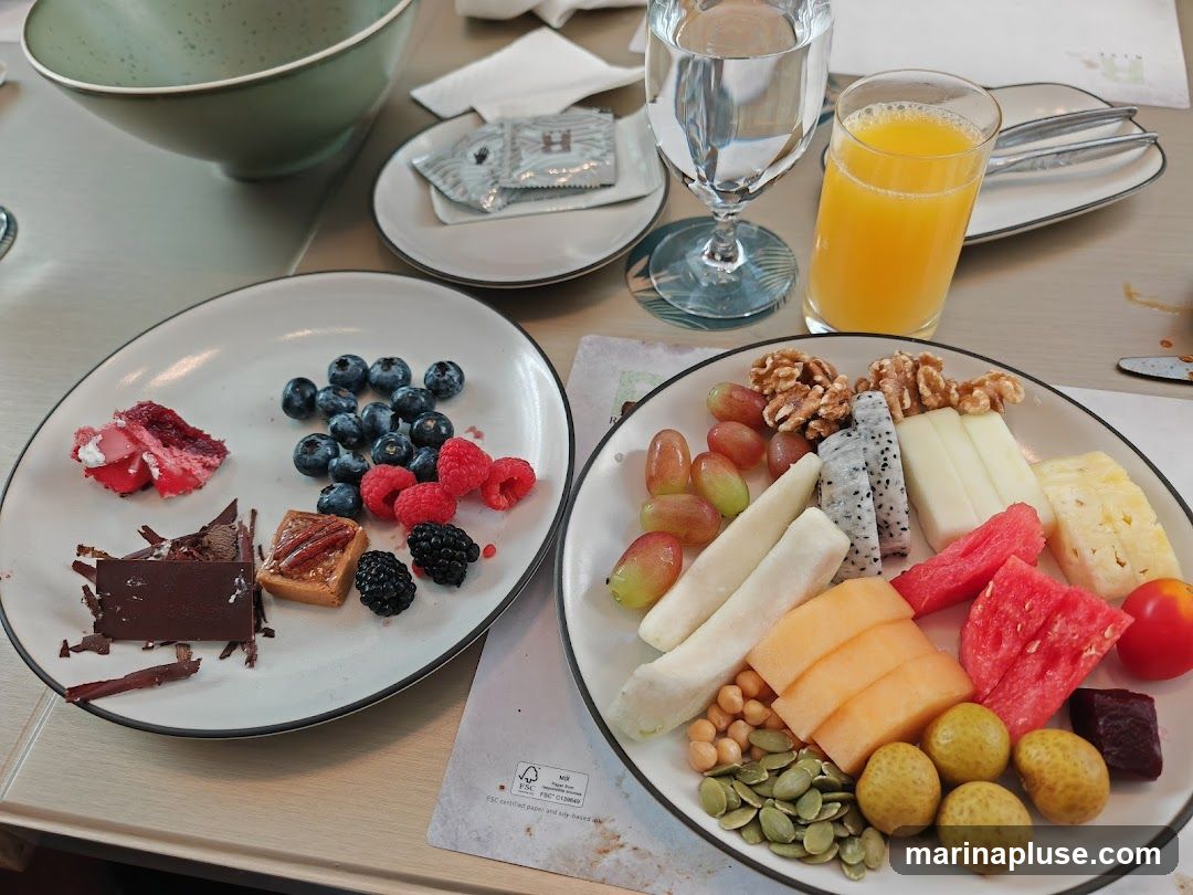 A close-up shot of two plates filled with various desserts and fruits, set on a table. The plate on the left contains a slice of red cake, blueberries, raspberries, blackberries, a small pastry, and chocolate shavings. The plate on the right features a selection of fruits including grapes, dragon fruit, melon, pineapple, watermelon, and a small red fruit, along with walnuts, chickpeas, pumpkin seeds, and small yellow fruits. A glass of orange juice and a glass of water are also visible, along with cutlery and a napkin.