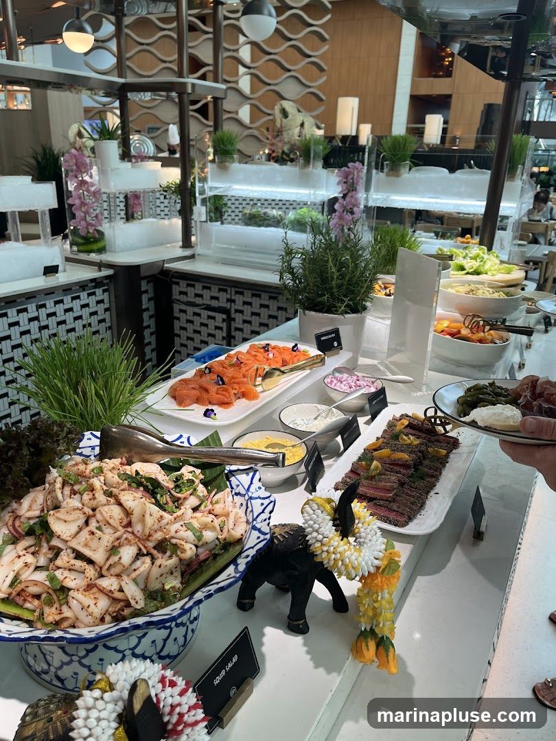 A buffet table laden with various dishes, including a large bowl of squid salad, sliced smoked salmon, a platter of sliced beef, and a salad bar with lettuce and tomatoes. Decorative elements like a small elephant statue and floral garlands are also visible.