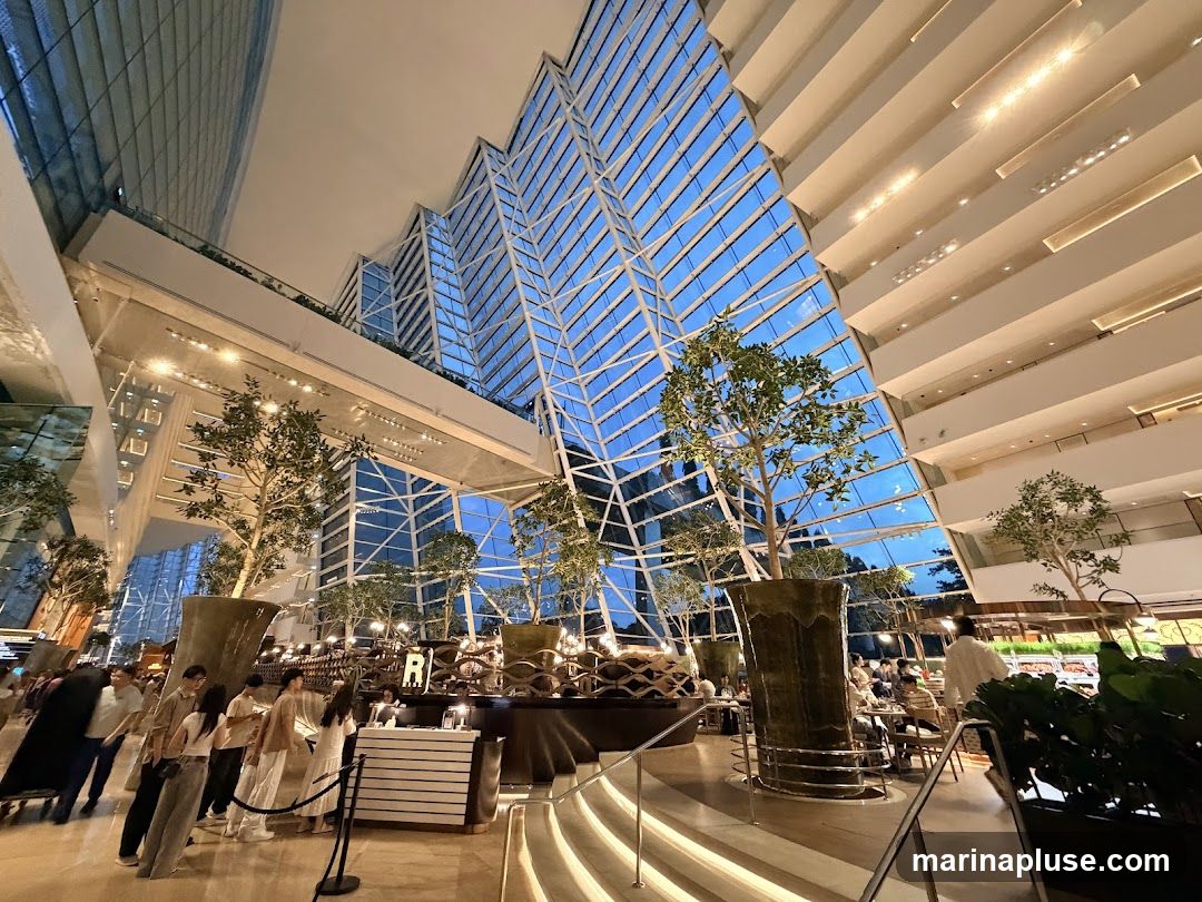A wide-angle shot of a spacious, modern atrium with a striking geometric glass ceiling. Tall, artificial trees in large planters are strategically placed throughout the space, adding a touch of nature. Multiple levels of balconies overlook the central area, and people are seen mingling and dining. The overall ambiance is sophisticated and airy, with natural light filtering through the glass structure.