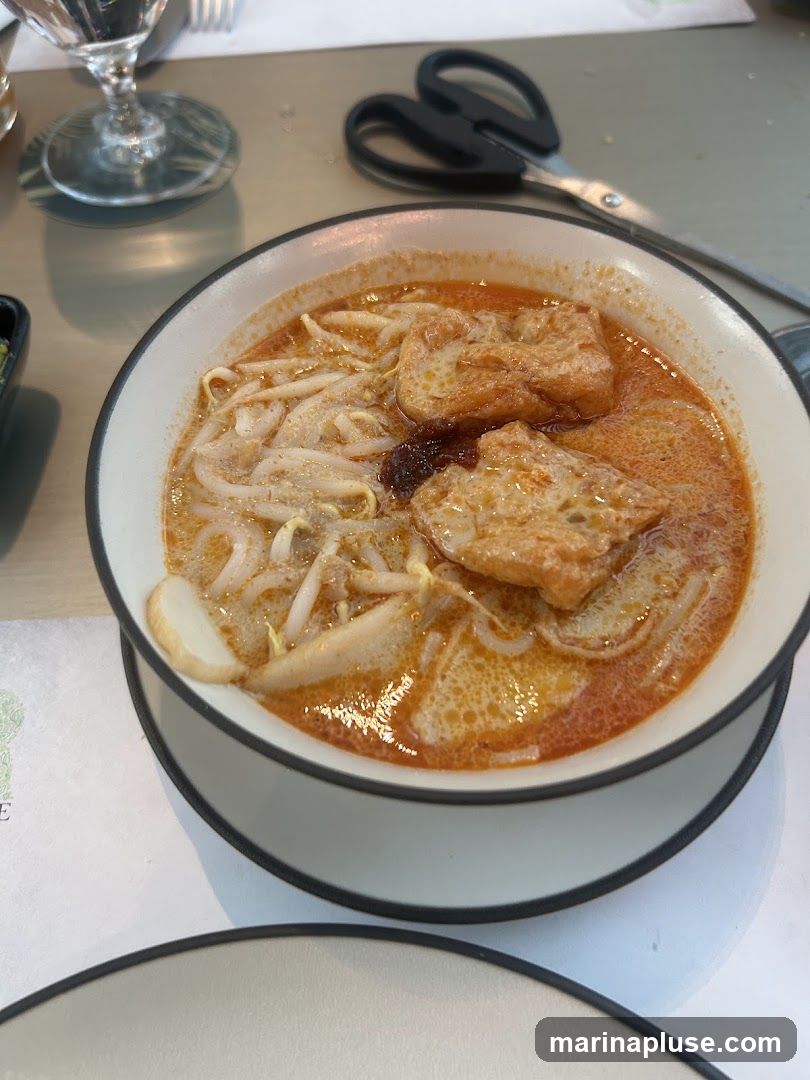 A close-up shot of a bowl of laksa noodle soup, a popular Southeast Asian dish. The soup is a rich, orange-colored broth, filled with noodles, bean sprouts, fried tofu, and a slice of fish cake. A dollop of chili paste is visible in the center of the bowl. In the background, a pair of black scissors rests on a metallic surface, along with a glass and a placemat.