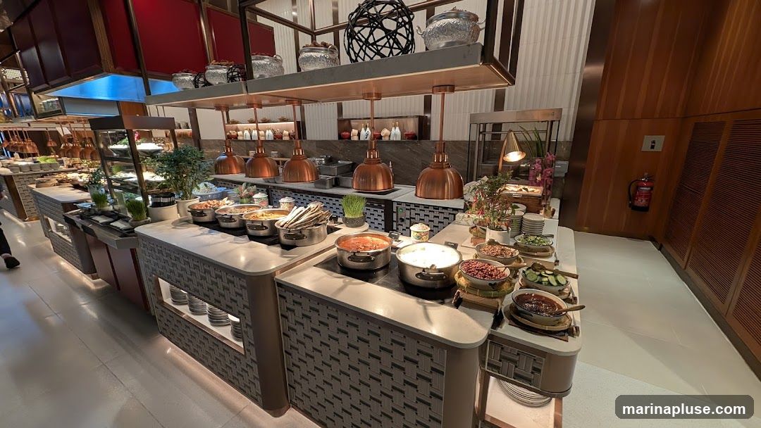 A wide shot of a buffet food display with multiple serving stations. Various dishes are presented in chafing dishes and bowls, including what appears to be rice, stews, salads, and side dishes. Overhead copper lamps provide warm lighting. The buffet area is decorated with plants and decorative items. A fire extinguisher is visible on a wooden paneled wall in the background.