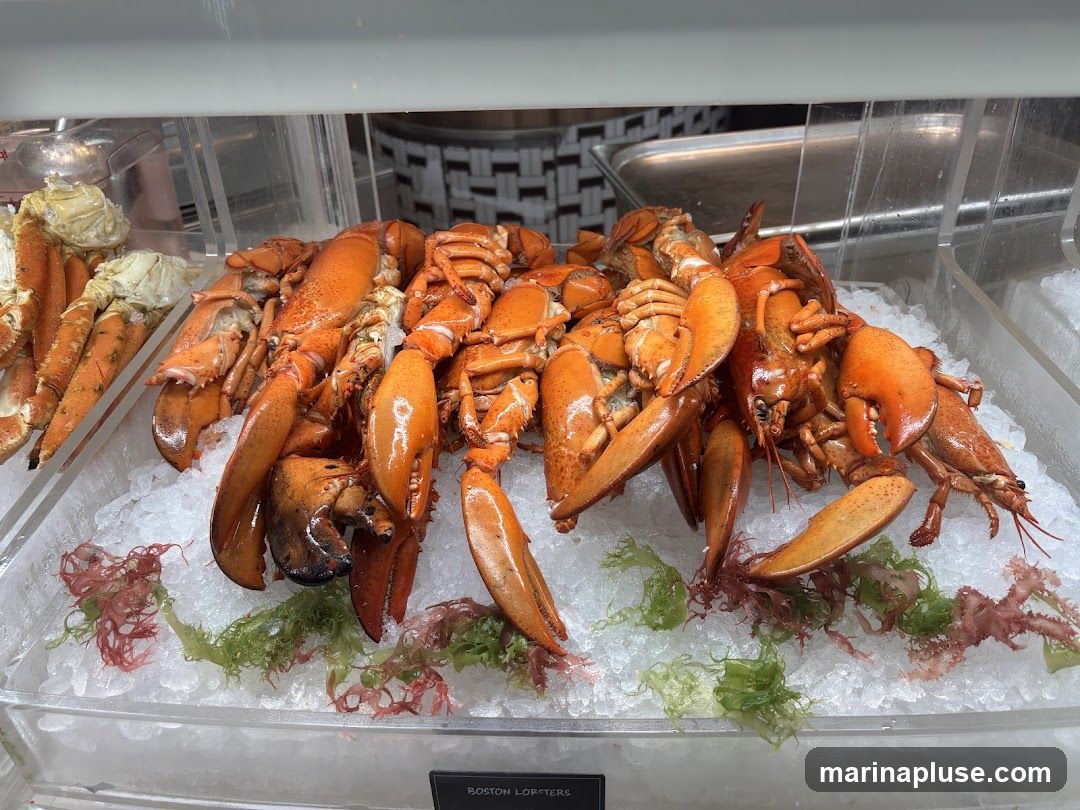 A display of cooked Boston lobsters and crab legs on a bed of crushed ice, garnished with seaweed. The lobsters are bright orange and piled together, with their claws prominently visible. Crab legs are arranged to the left of the lobsters. A small black label at the bottom reads "BOSTON LOBSTERS" but no price is visible.